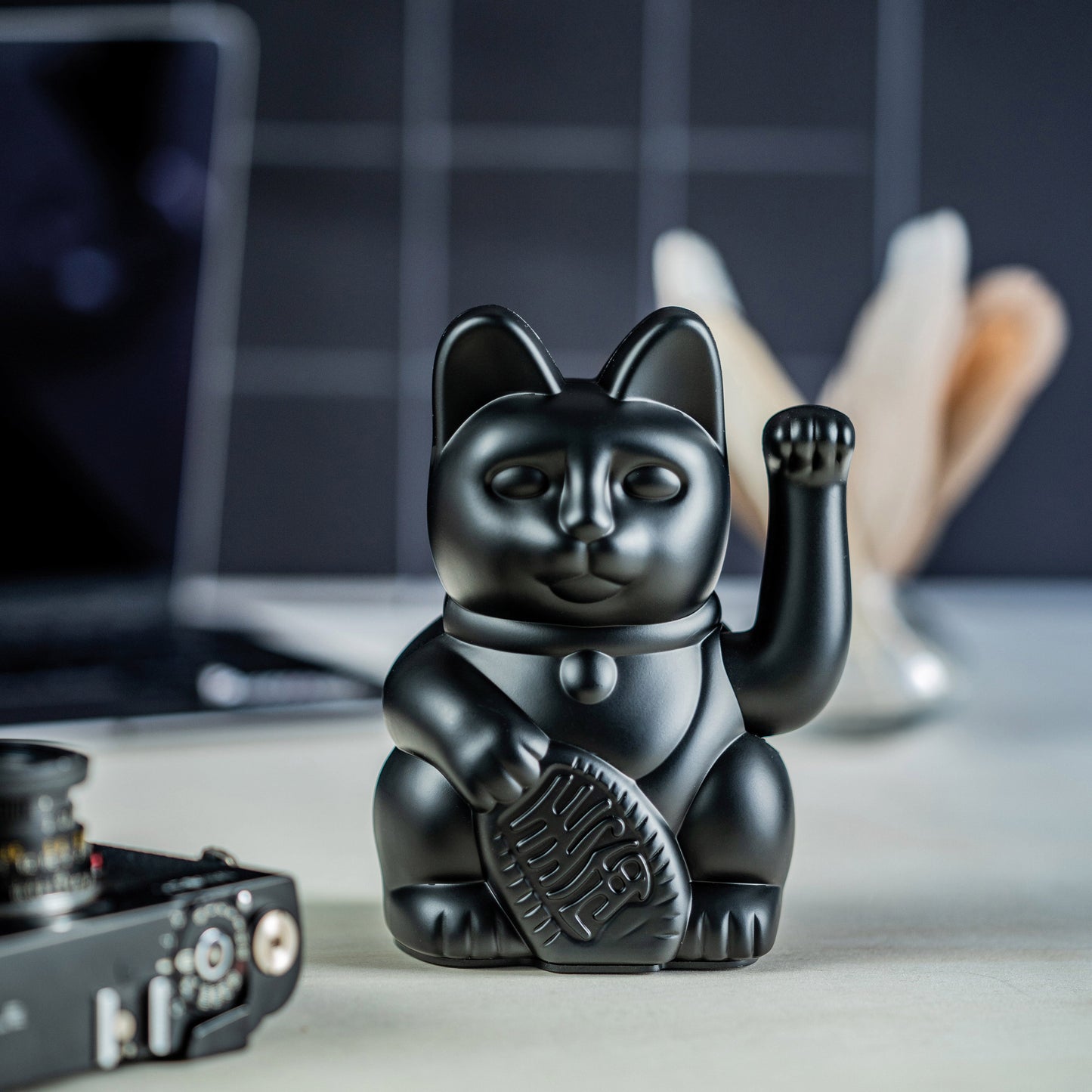 Matte schwarze Winkekatze „Lucky Cat“ von Donkey in der Größe Mini auf einem Tisch arrangiert, mit Black-Vibe-Deko sowie teilweise sichtbarer Kamera und Computer im Hintergrund.