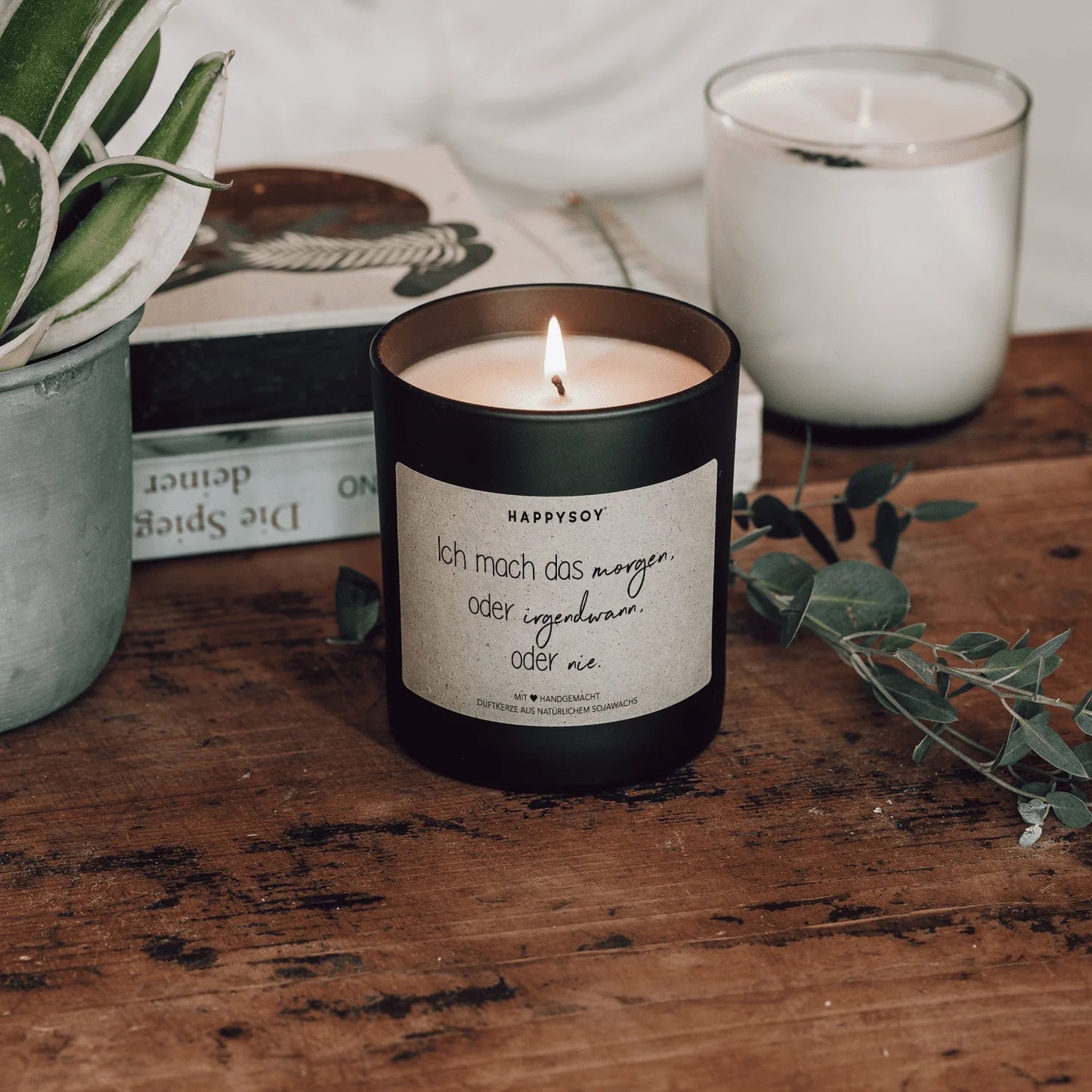 Angezündete schwarze Sojawachskerze von HappySoy mit Spruch ‘Ich mach das morgen, oder irgendwann, oder nie.’, dekorativ aufgestellt neben Büchern und Pflanzen