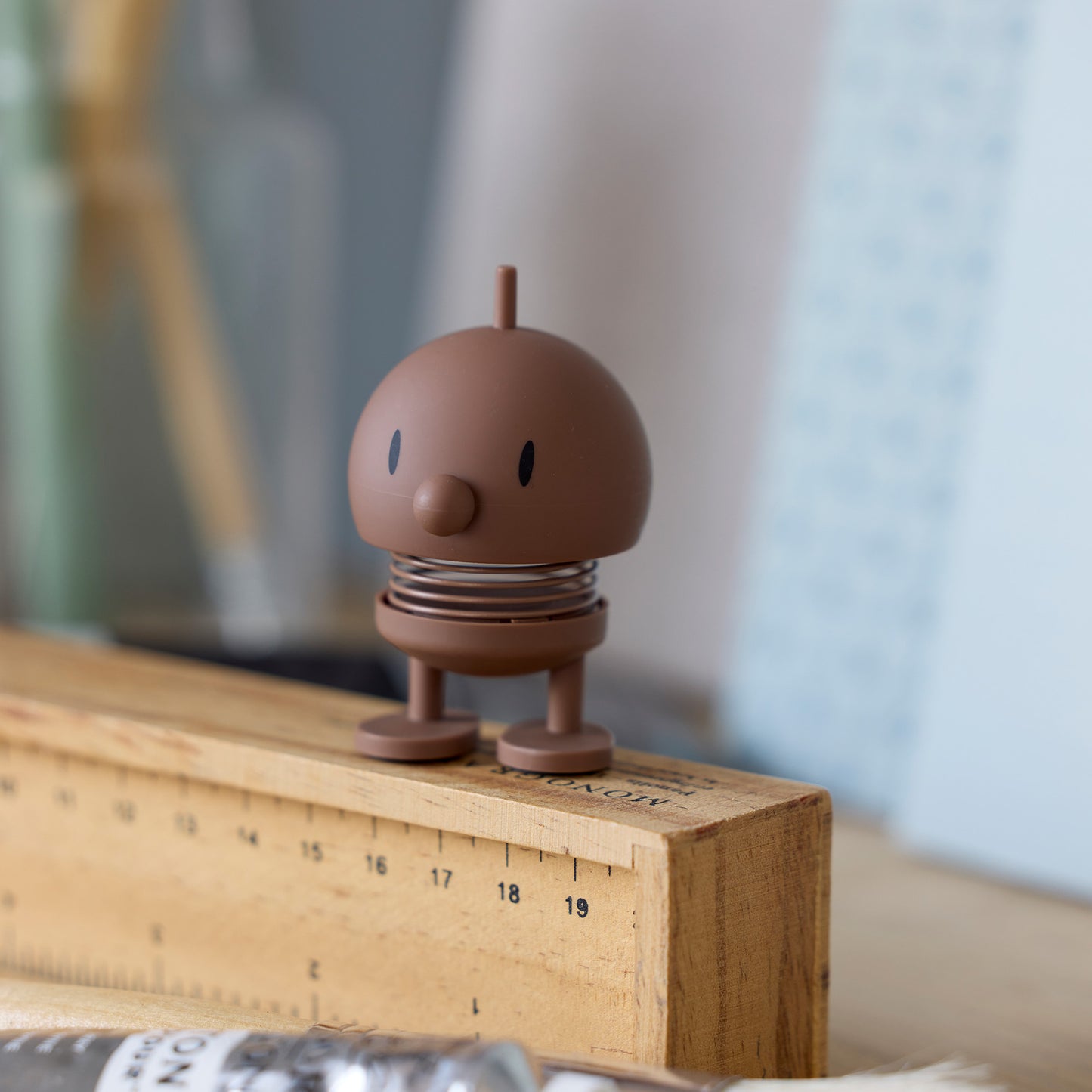 Mini Hoptimist Bumble in Soft-Optik in Schokobraun steht auf einem Holzlineal, mit runder Form und sichtbarer Feder als Wackelmechanismus