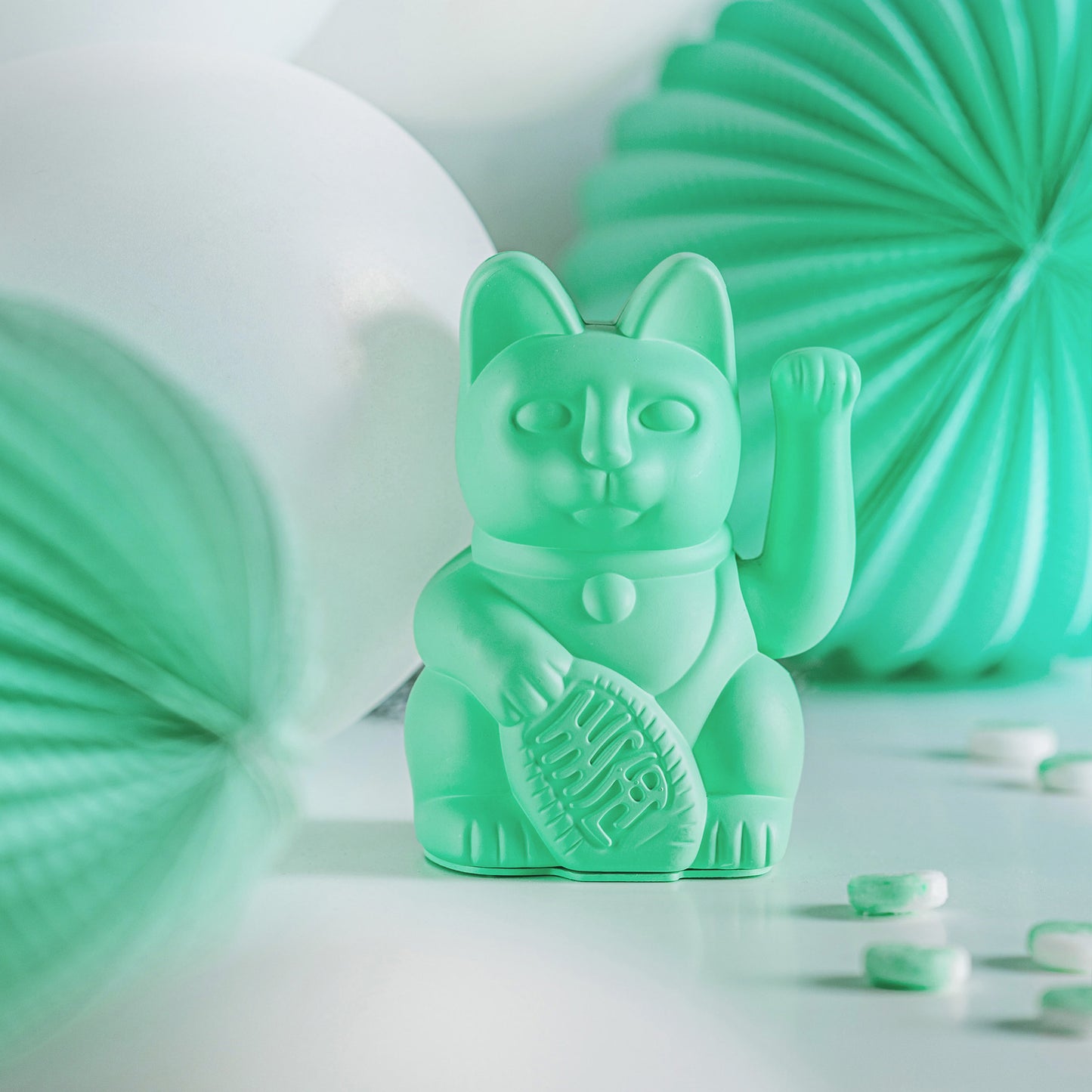 Mintgrüne Mini Winkekatze „Lucky Cat“ von Donkey auf einem Tisch mit mintgrünen Papier-Dekoballons und Bonbons in Weiß und Mint arrangiert.