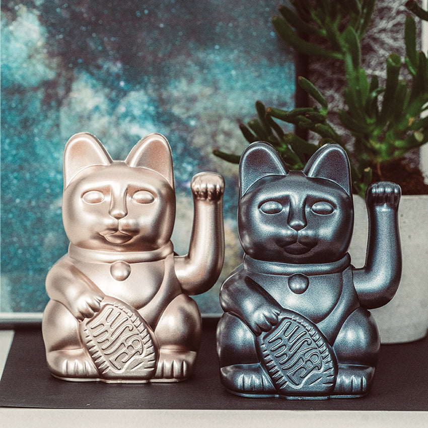 Moonlight Winkekatze von Donkey in der Variante Moonlight neben der Galaxy-Variante. Die beiden dekorativen Glückskatzen sind auf einem Tisch arrangiert und werden von stilvoller Deko und passenden Galaxy-Motiven im Hintergrund ergänzt, wodurch ein moderner, harmonischer Kontrast entsteht.