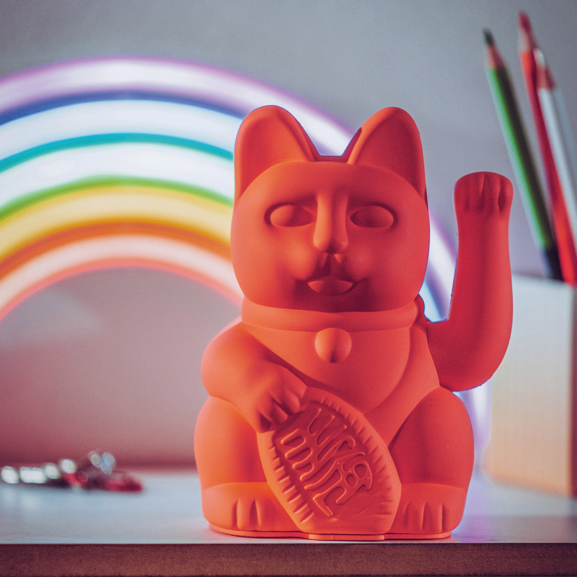 Neonorange Winkekatze „Lucky Cat“ von Donkey in der Größe Regular vor einem Regenbogen-LED-Schild im Old-School-Stil arrangiert.