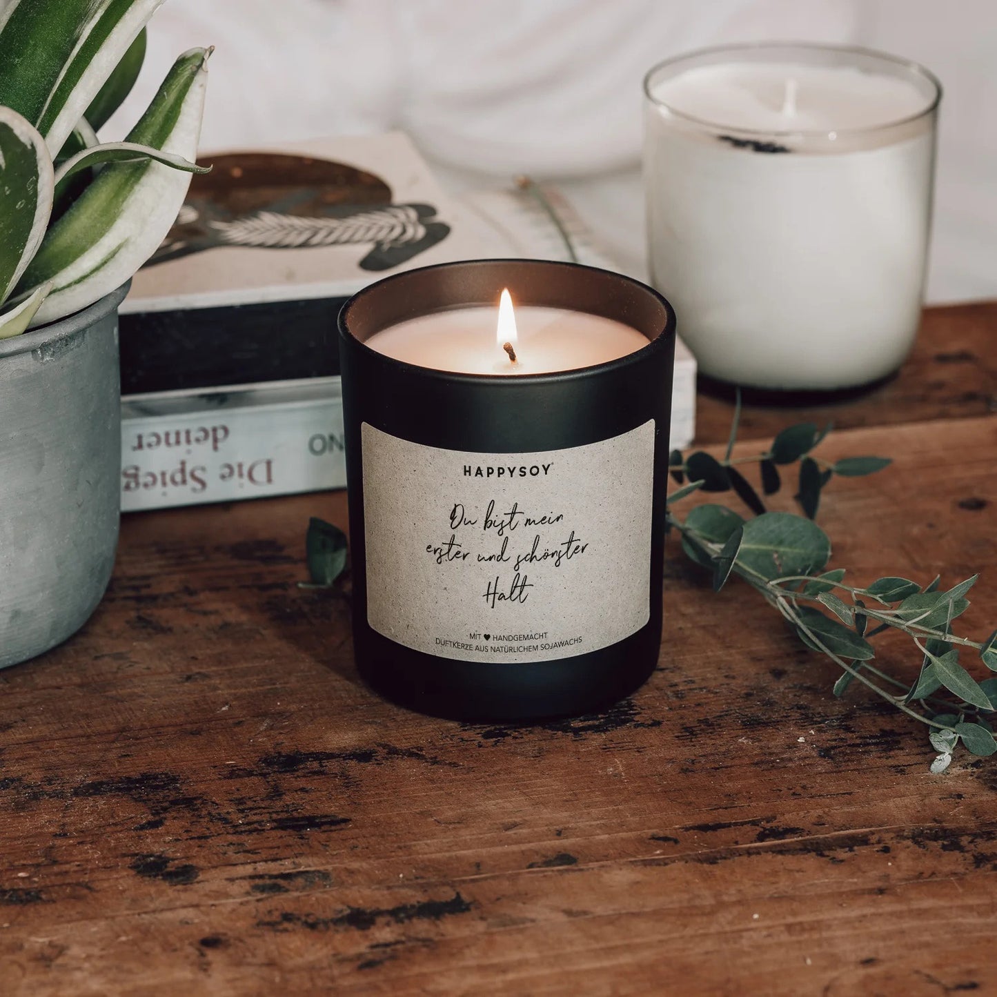 Angezündete schwarze Sojawachskerze von HappySoy mit Spruch ‘Du bist mein erster und schönster Halt’, dekorativ aufgestellt neben Büchern und Pflanzen.