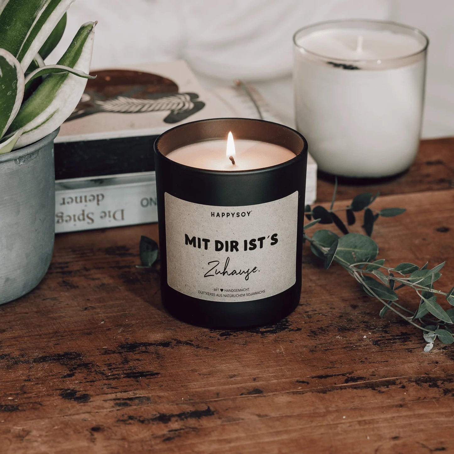 Schwarze Sojawachskerze von HappySoy mit Spruch ‘Mit dir ist's zuhause’, angezündet und dekorativ aufgestellt neben Büchern und Pflanzen