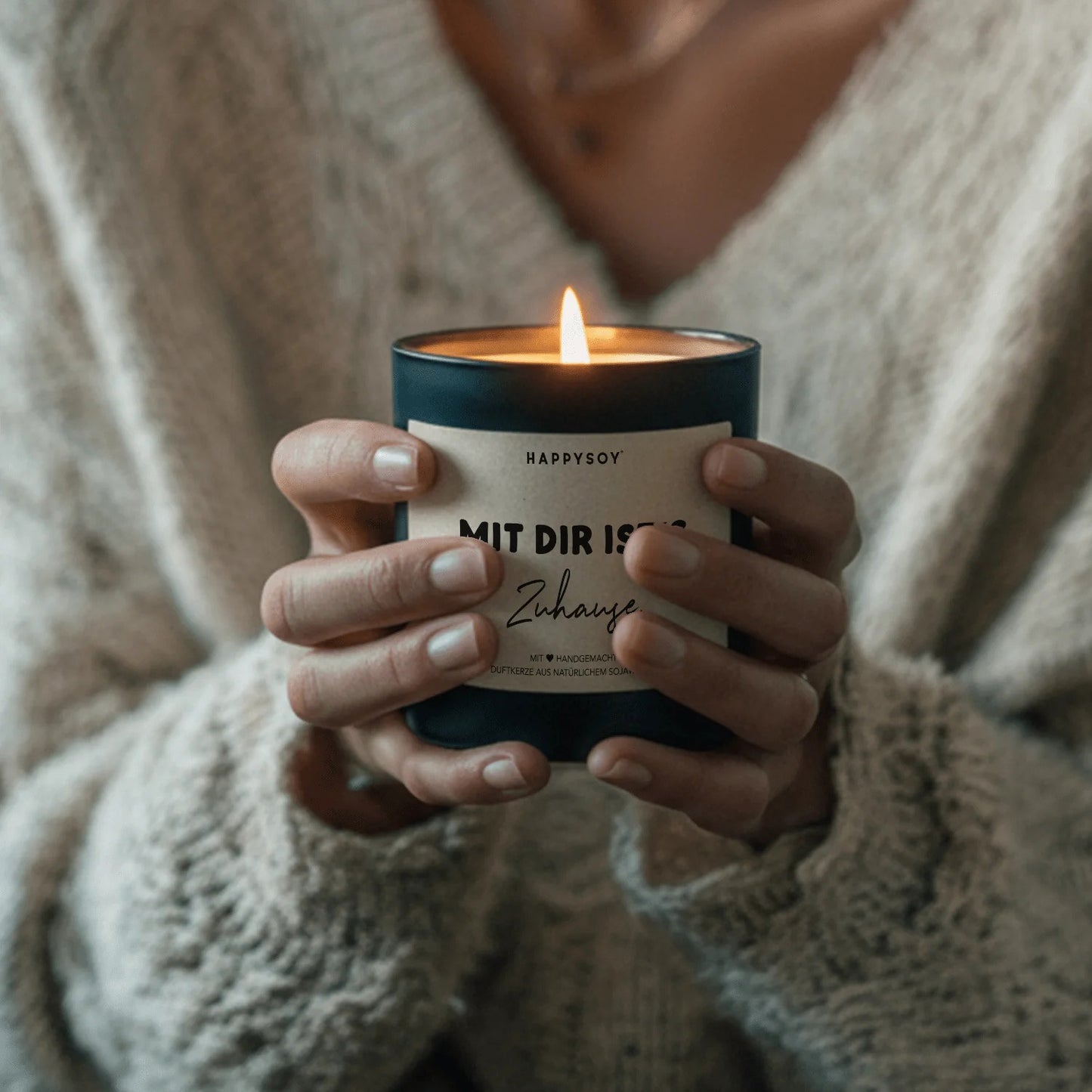 Jemand hält eine angezündete schwarze Sojawachskerze von HappySoy mit Spruch ‘Mit dir ist's zuhause’ in beiden Händen.
