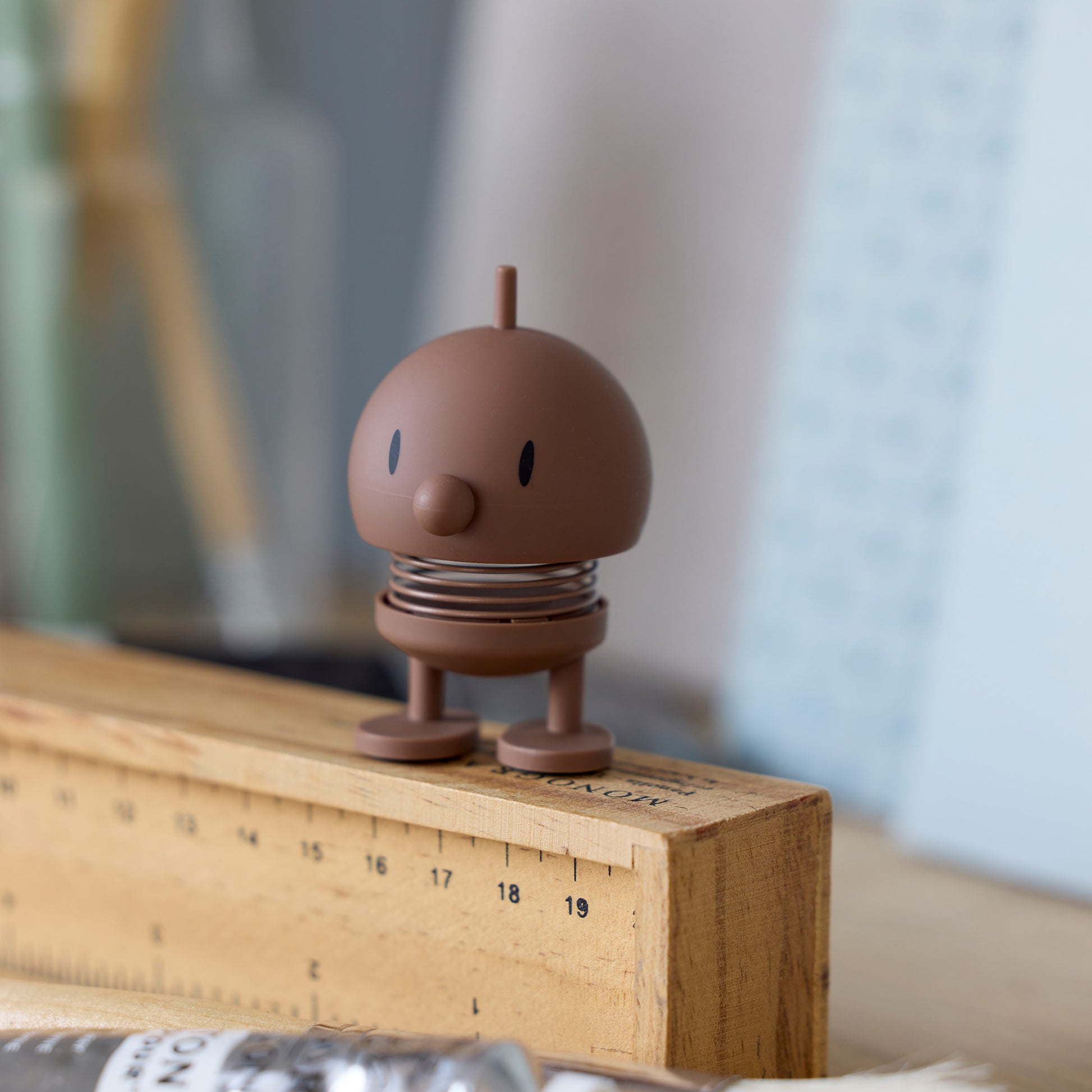 Mini Hoptimist Bumble in Soft-Optik in Schokobraun steht auf einem Holzlineal, mit runder Form und sichtbarer Feder als Wackelmechanismus
