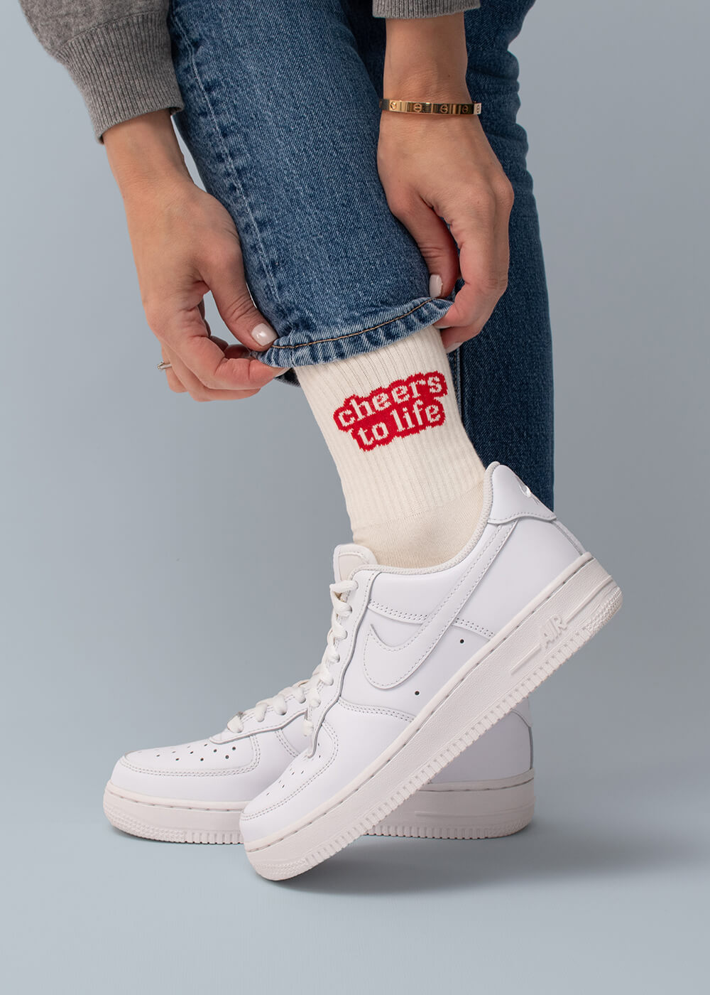 Weiße Crew-Socken mit rotem Schriftzug „Cheers to Life“, getragen mit weißen Sneakers und Jeans