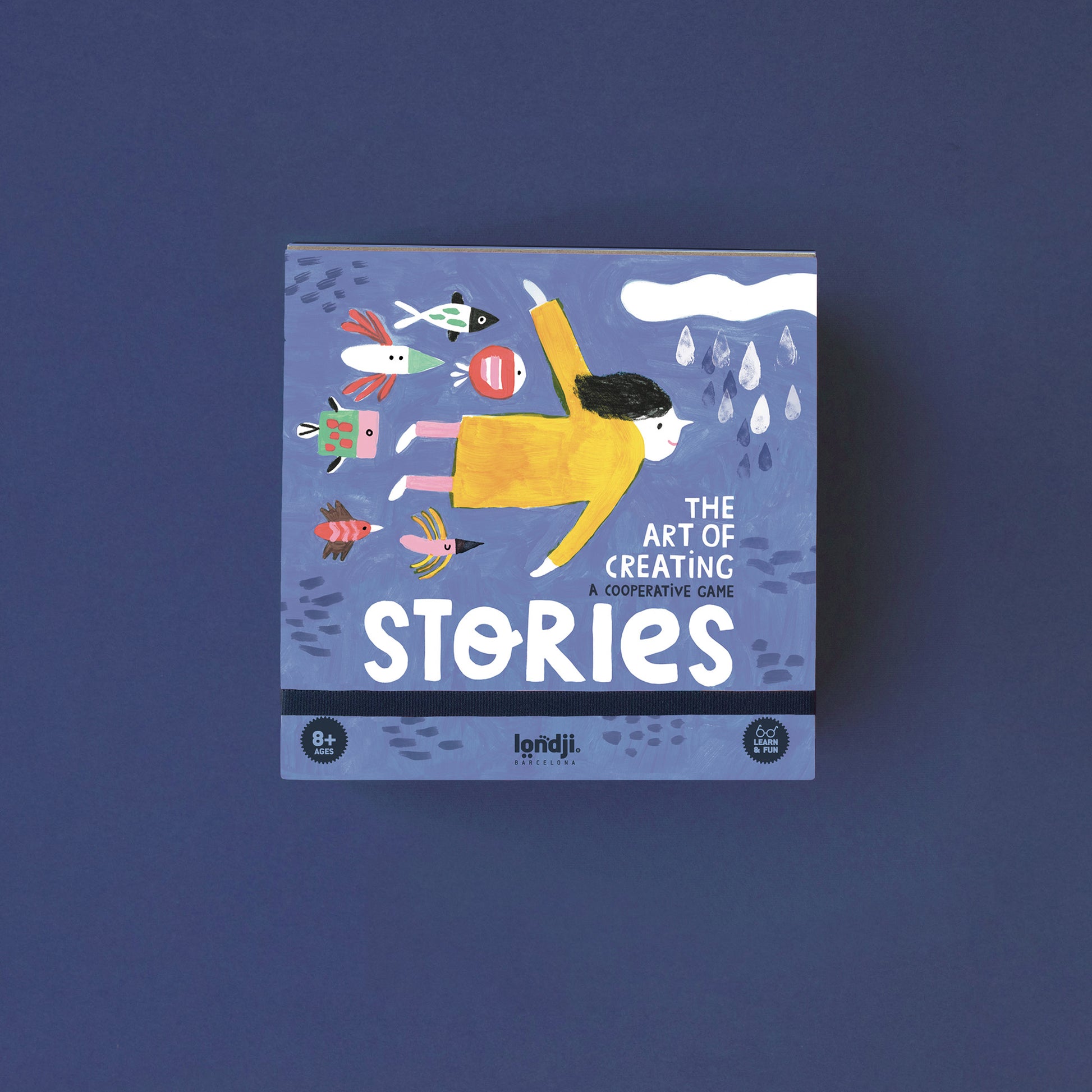 Spiel „Stories“ von Londji, blaue Spielbox mit Illustration eines Mädchens im gelben Kleid im Wasser, umgeben von Fischen