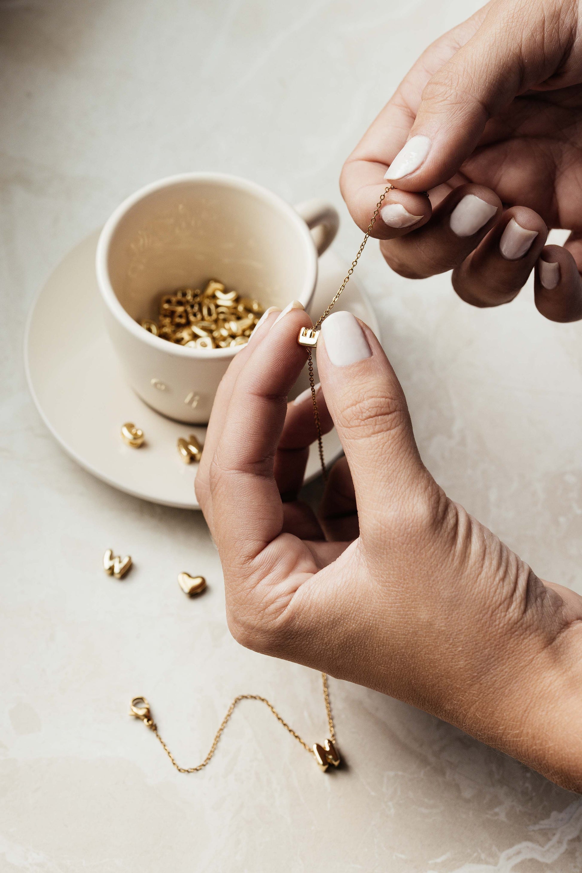 Teetasse gefüllt mit goldenen Bubble-Letter-Charms, während ein Buchstabe E auf eine Kette aufgefädelt wird