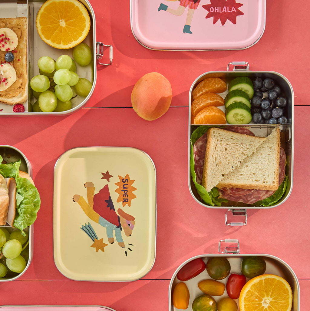 Zwei Edelstahl-Lunchboxen mit gelbem und rosa Deckel, mit Superhelden-Motiven, geöffnet und mit Snacks gefüllt, auf einer Picknickdecke als Moodbild
