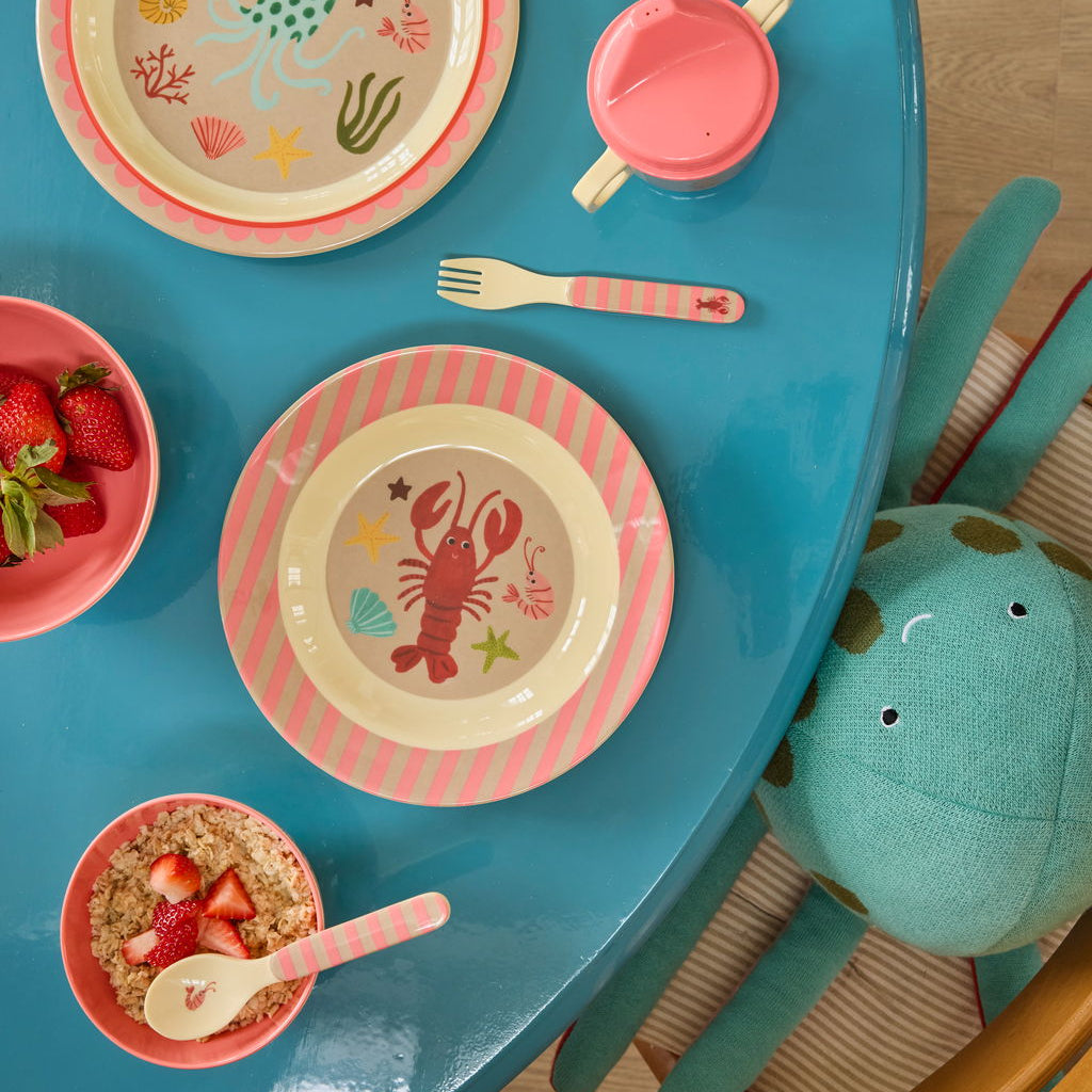 Moodbild mit rosa Kinderbesteck zusammen mit großem und kleinem rundem Kinderteller sowie passender Baby-Trinkflasche, harmonisch aufeinander abgestimmt und liebevoll arrangiert auf dem Tisch