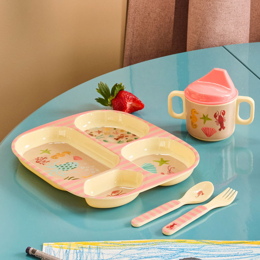 Stimmungsbild des rosafarbenen Kinderteller mit Abtrennungen zusammen mit passendem Besteck und Babyflasche, liebevoll arrangiert auf einem Tisch in sanften, harmonischen Farben